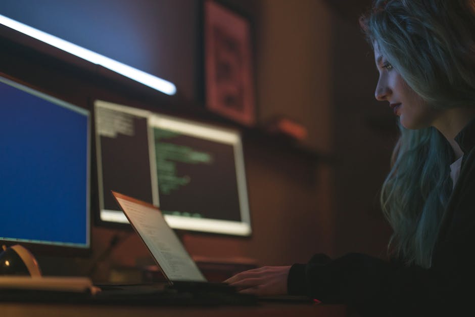 Person at a home desk with multiple monitors showing code and documentation, coffee mug nearby, books stacked beside keyboard, late night warm lighting, self-directed learner energy