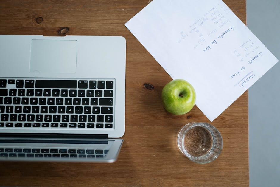 A minimalist writing setup with just a laptop, a glass of water, and a notepad on a plain wooden desk, natural window light, overhead view