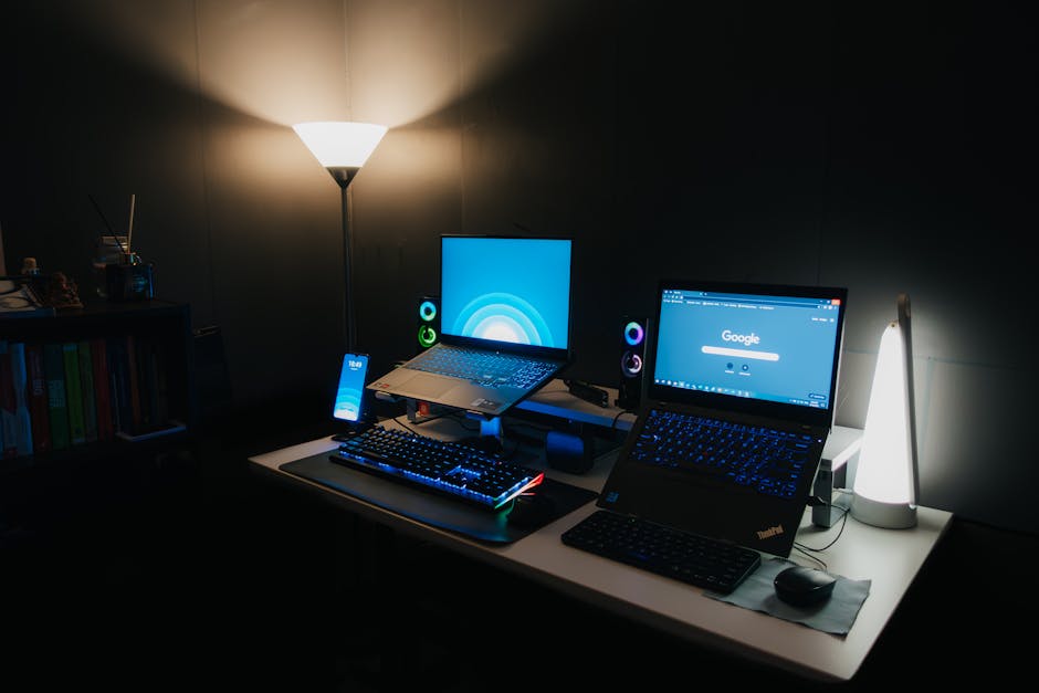 A cluttered but functional home desk setup with an old laptop, a mini PC, and networking gear — warm lighting, casual and real