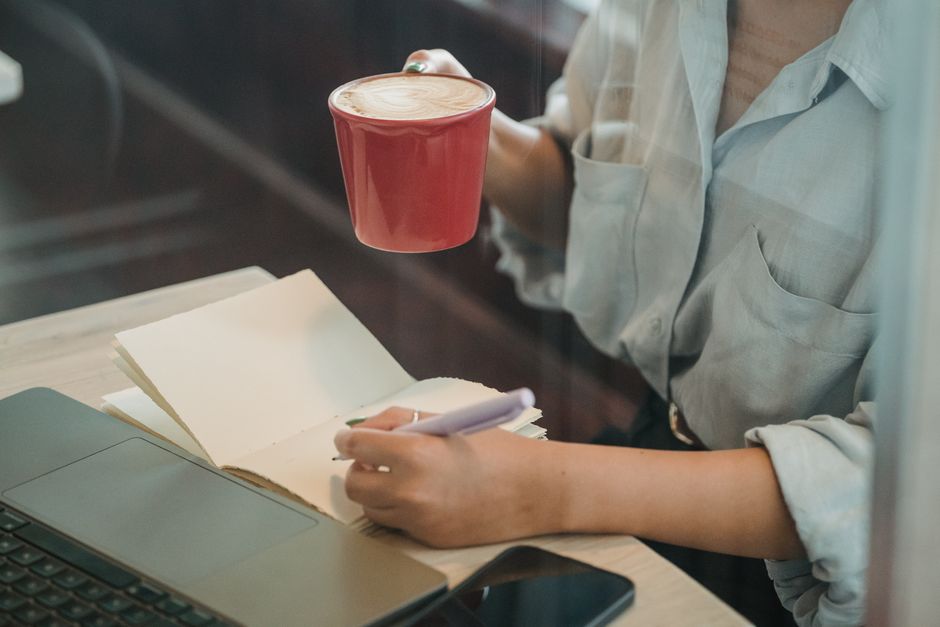 Person sitting at a desk with a laptop, handwritten budget notebook, and a cup of coffee, focused and calm, warm natural light, candid documentary style