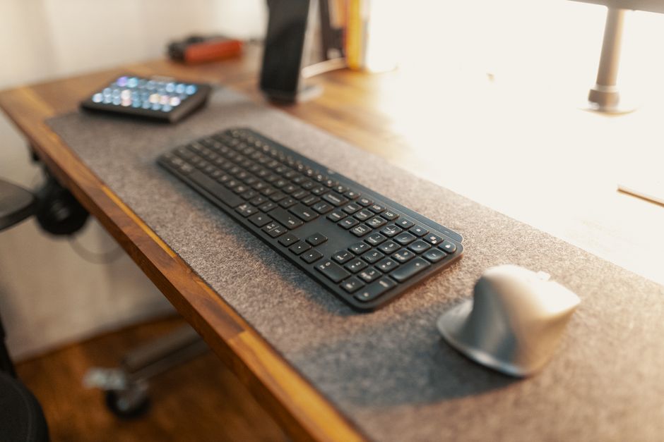 Close-up of a mechanical keyboard, external monitor stand, and ergonomic mouse on a wooden desk — professional workspace, warm lighting